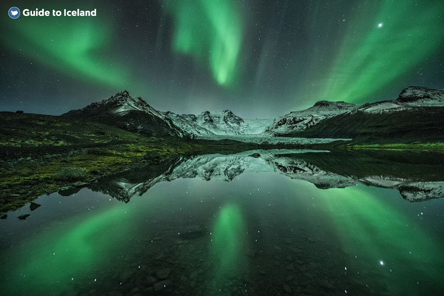 北极光把天空描绘成鲜艳的绿色，山脉和灯光反射到下面纯净的水上。