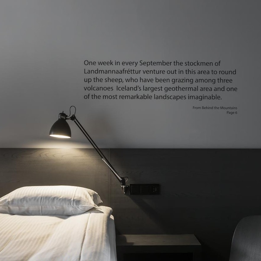 A lamp shines over a comfortable bed, with a book excerpt on the wall at Skuggi Hotel Reykjavík by Keahotels. 