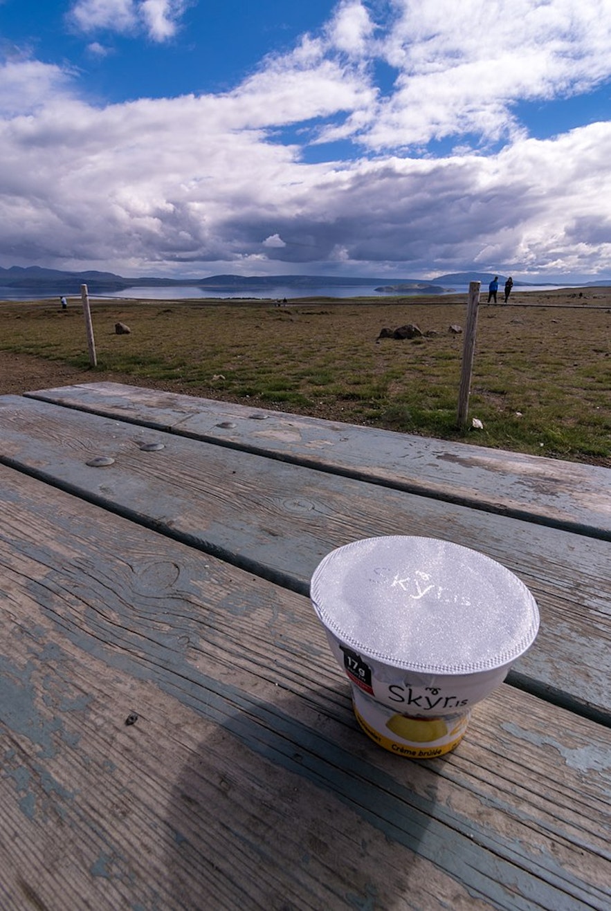 A closed tub of skyr with a foil lid on a picnic bench outside, with mountains and a fjord in the background.