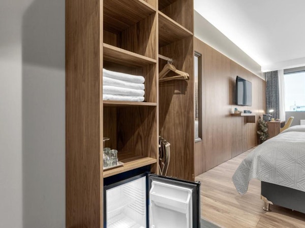 A wooden closet with coat hangers on one side and folded towels, a fridge, and drinking glasses on the other side.