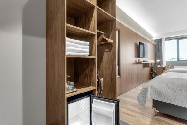 A wooden closet with coat hangers on one side and folded towels, a fridge, and drinking glasses on the other side.