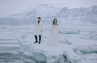 A couple wearing wedding outfits pose for a photo on a glacier lagoon iceberg during a three-day honeymoon photography tour on Iceland’s South Coast.