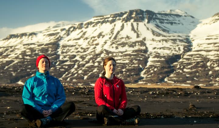 Two people sit cross-legged on a black sand beach in front of snowy mountains with their eyes closed and practice mindfulness meditation.