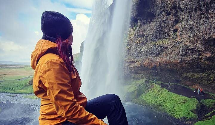 A person in a waterproof jacket and beanie hat sits and looks at a waterfall on Iceland's South Coast.