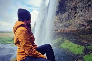 A person in a waterproof jacket and beanie hat sits and looks at a waterfall on Iceland's South Coast.
