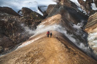 Two people walk through the Hveradalir geothermal valley in the Icelandic Highlands, with steam rising around the peaks and snowy terrain stretched out behind.