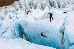 A person stands, harnessed up at the top of an ice wall, while another is lowered into a deep blue crevasse.