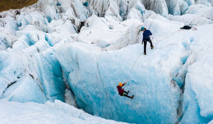 A person stands, harnessed up at the top of an ice wall, while another is lowered into a deep blue crevasse.