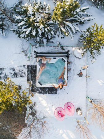 A birdseye view of two people relaxing in a jacuzzi at Kalda Lyngholt amid a snow-covered landscape.