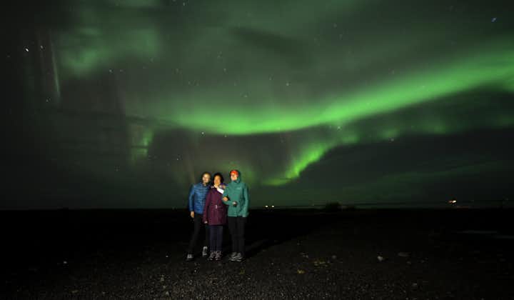 Three people stand together watching the northern lights over the South Coast of Iceland.