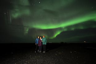 Three people stand together watching the northern lights over the South Coast of Iceland.