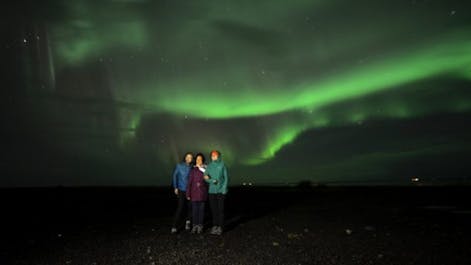 Three people stand together watching the northern lights in the sky above the South Coast of Iceland.