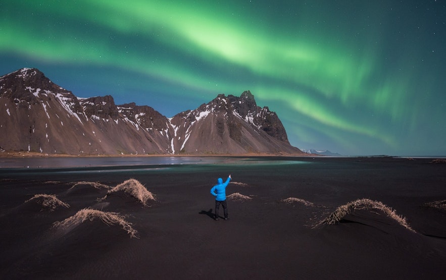 A person in a blue parka pointing up in the sky at the northern lights dancing over Vestrahorn Mountain in Iceland in winter