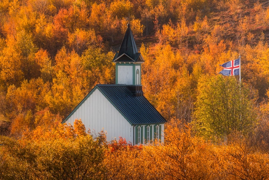 Die Kirche von Thingvellir umgeben von Herbstbäumen in Orange, Gelb und Rot mit der isländischen Flagge