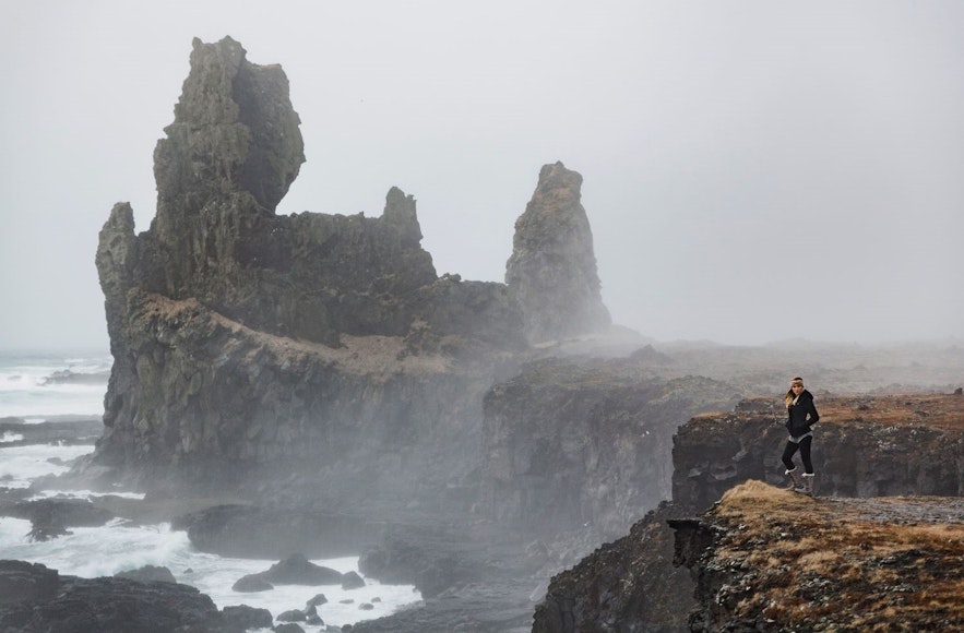 Klippen und Lavaformationen vor der Küste der Halbinsel Snaefellsnes in Island an einem sehr windigen Tag mit einer am Ufer stehenden Frau
