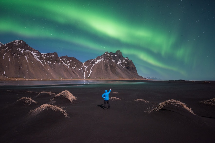 Person dressed in blue parka and black winter pants standing on black sand and pointing to the northern lights in the sky above Vestrahorn mountain
