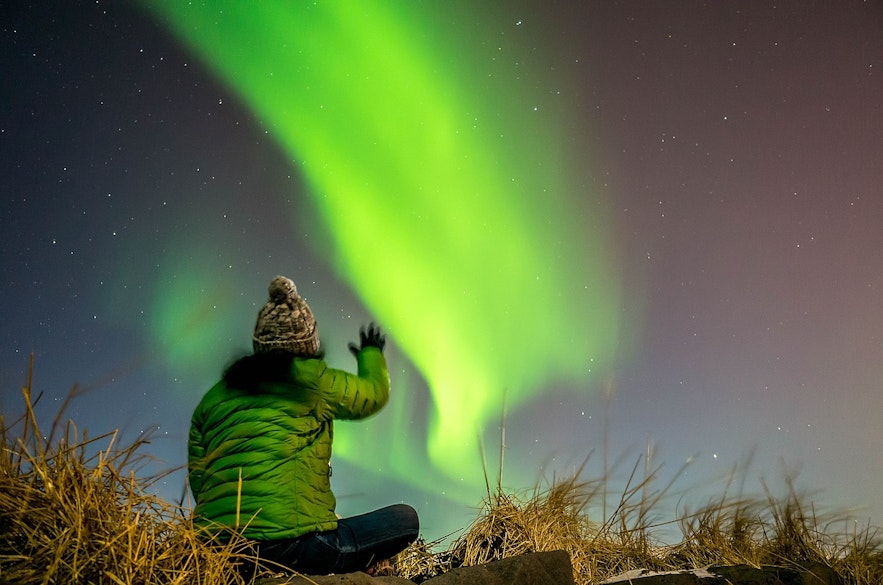 一个人坐在草地上，看着冰岛上空的北极光
